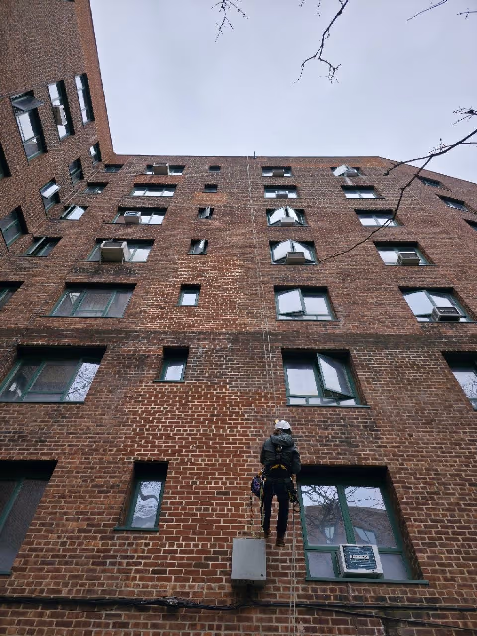 Worker in safety gear rappelling down a tall brick apartment building with multiple windows and air conditioners.