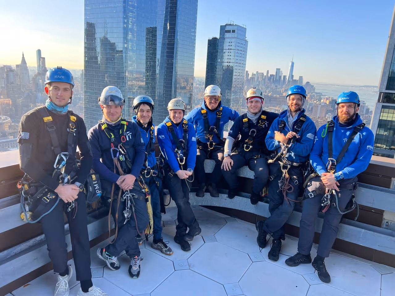 Group of eight workers wearing helmets and harnesses posing on a rooftop with city skyscrapers in the background during daylight.