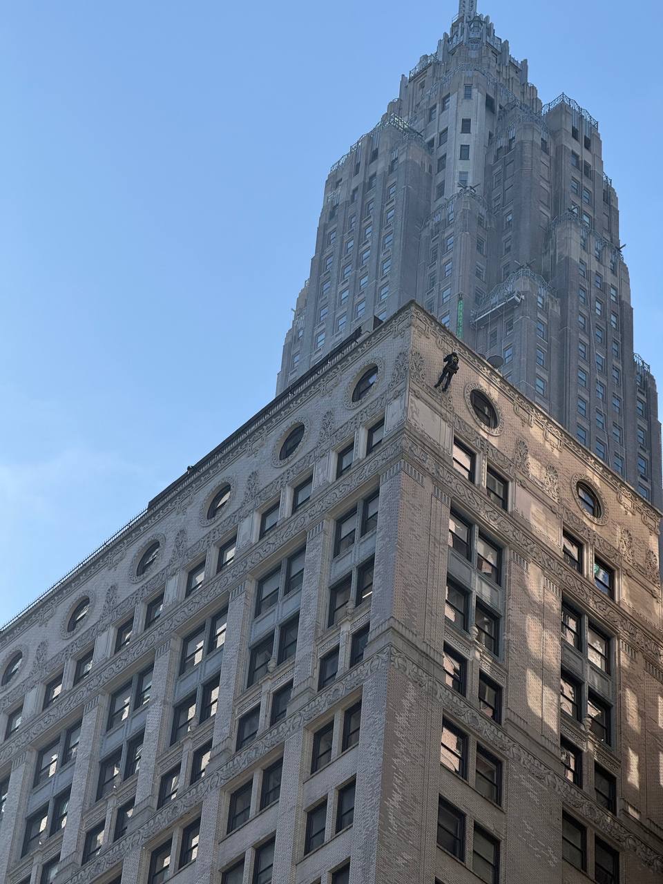 View looking up at a tall, historic skyscraper with detailed architecture and a worker suspended on the corner cleaning windows.