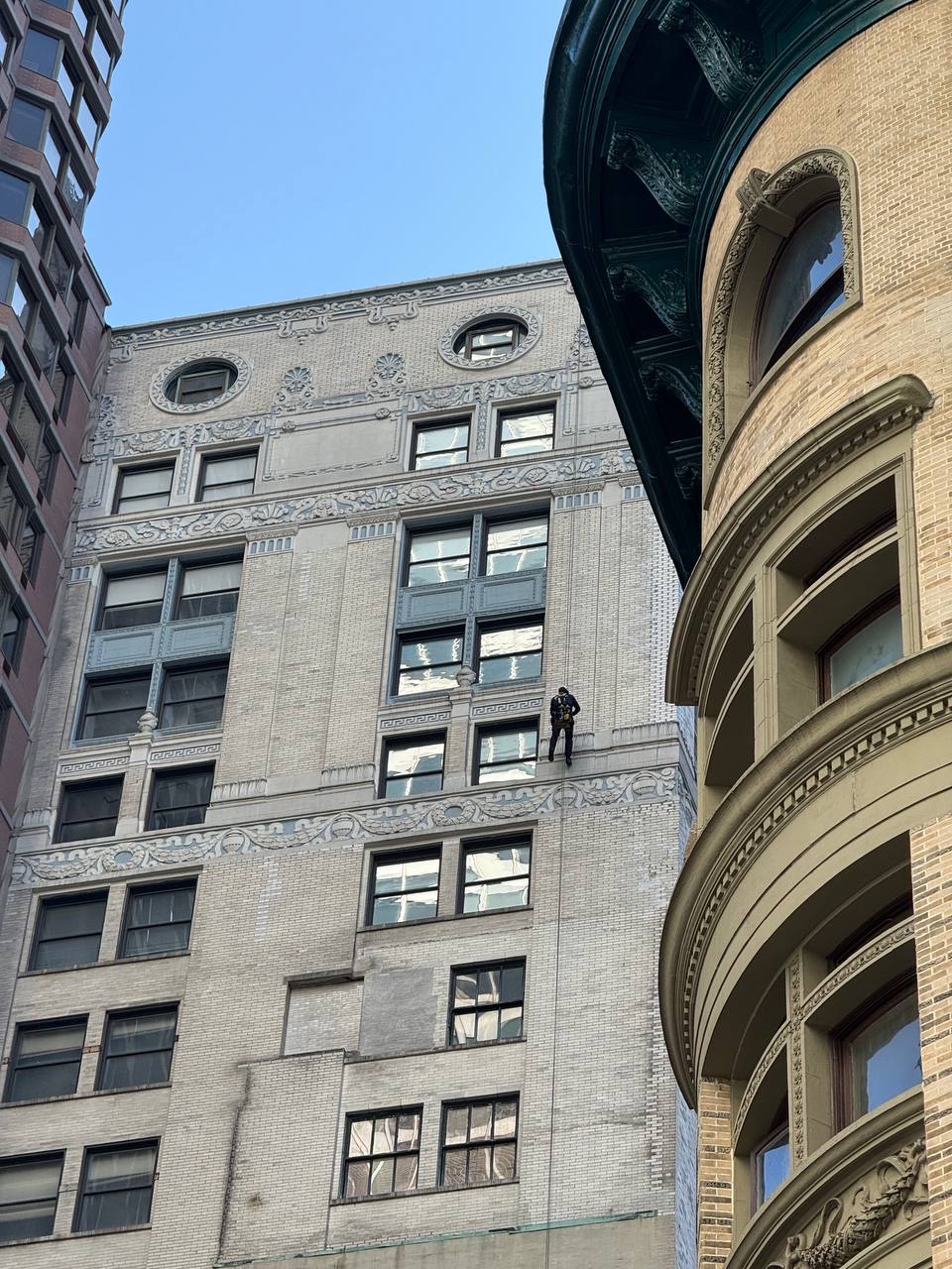 Window cleaner rappelling down a decorative tall building with large windows and ornate architectural details under clear blue sky.