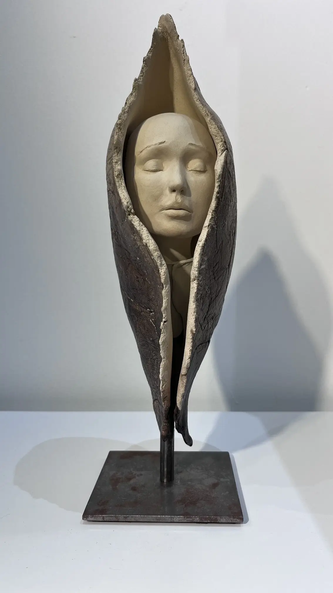 Sculpture en grès beige et gris sombre représentant un visage serein enveloppé dans une forme organique protectrice verticale.