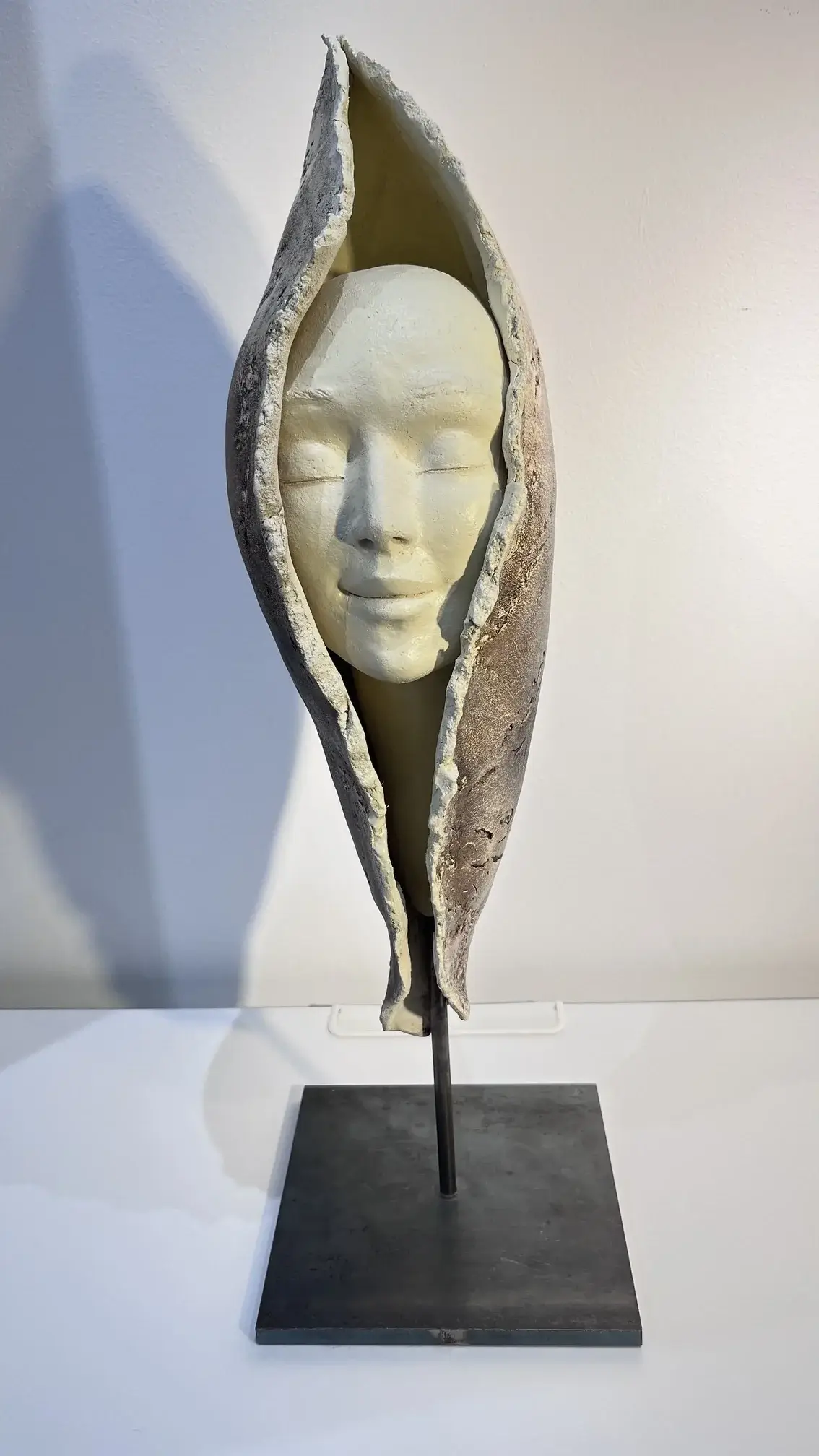 Sculpture en grès blanc et gris, visage souriant enveloppé dans une forme végétale protectrice sur socle en métal noir minimaliste.