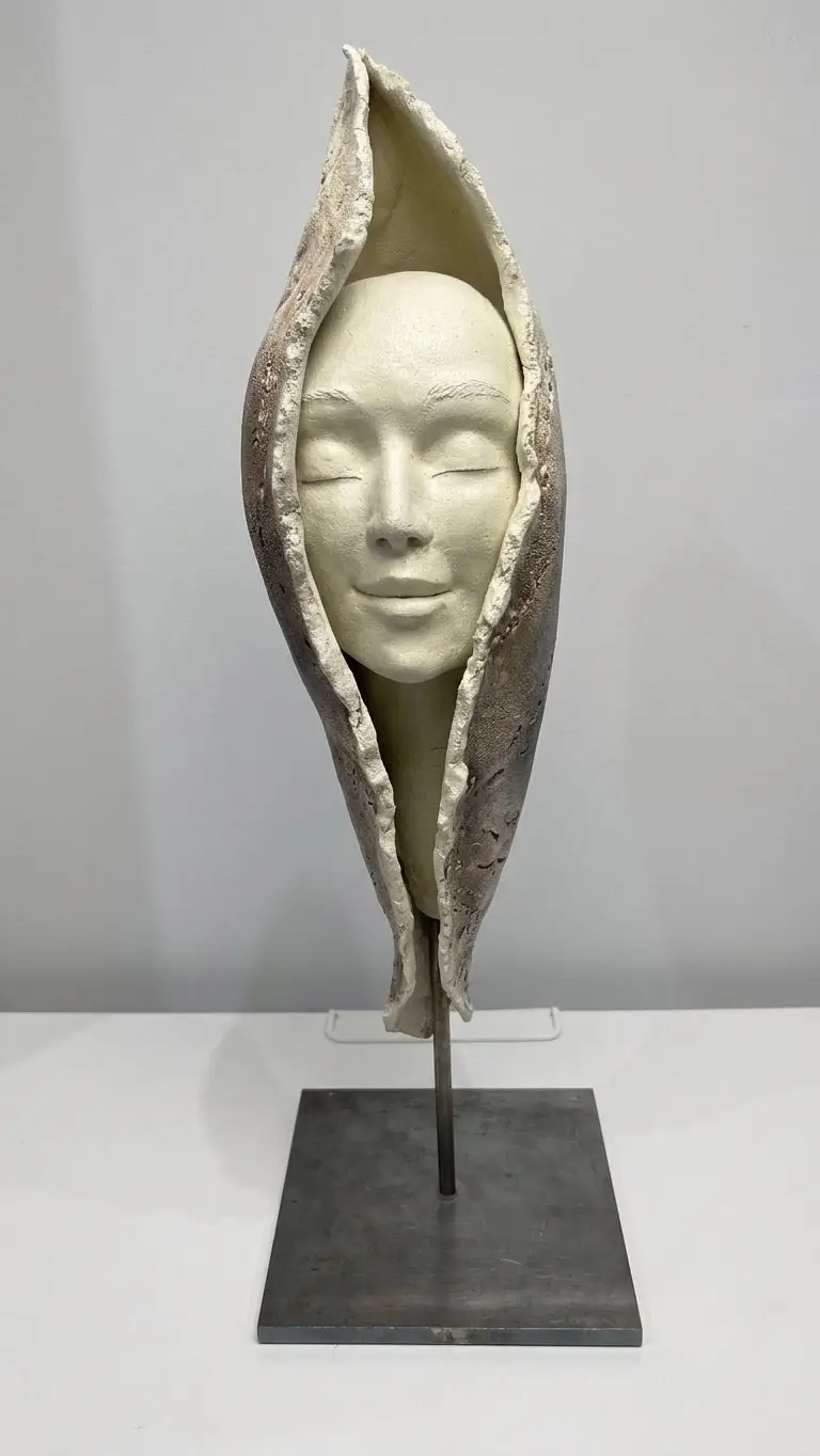 Sculpture en grès blanc et gris, visage souriant enveloppé dans une forme végétale protectrice sur socle en métal noir minimaliste.