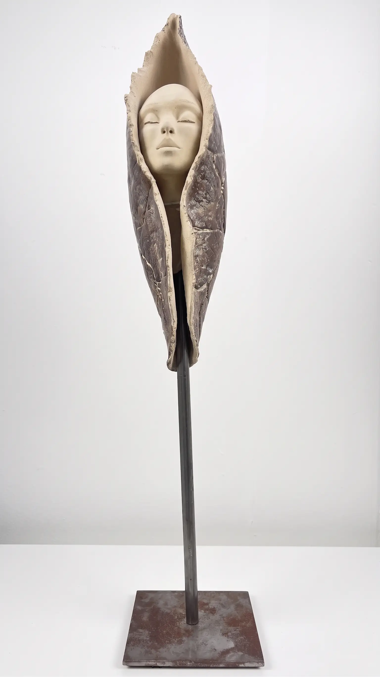 Sculpture en grès beige et brun sombre, visage apaisé enveloppé dans une forme organique verticale sur socle en métal noir.