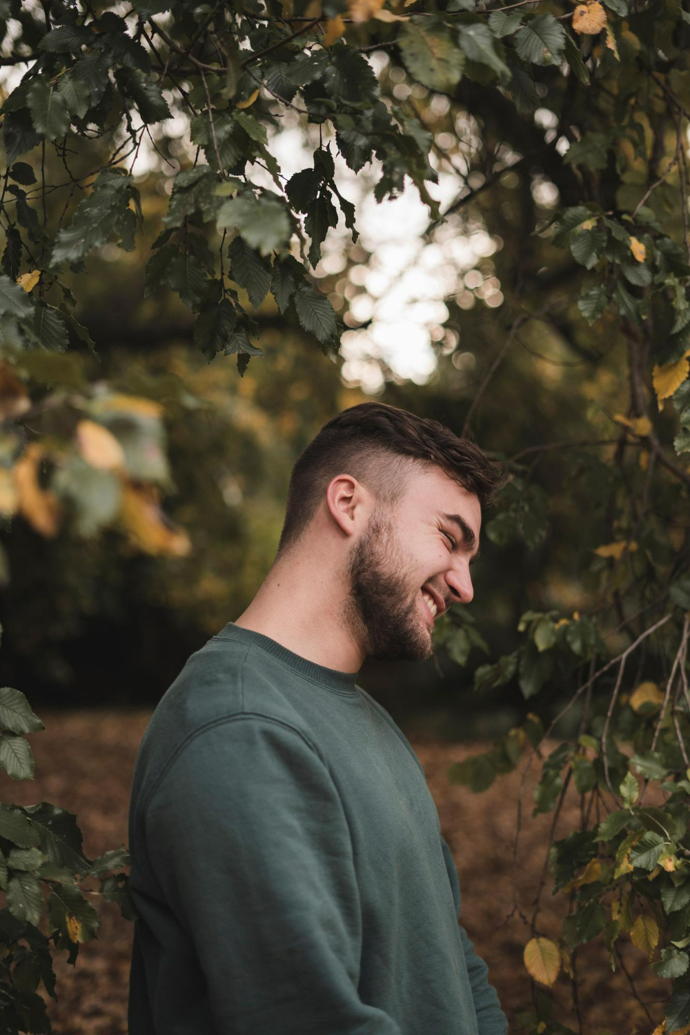 Young bearded man smiling with eyes closed among green and yellow leaves outdoors.