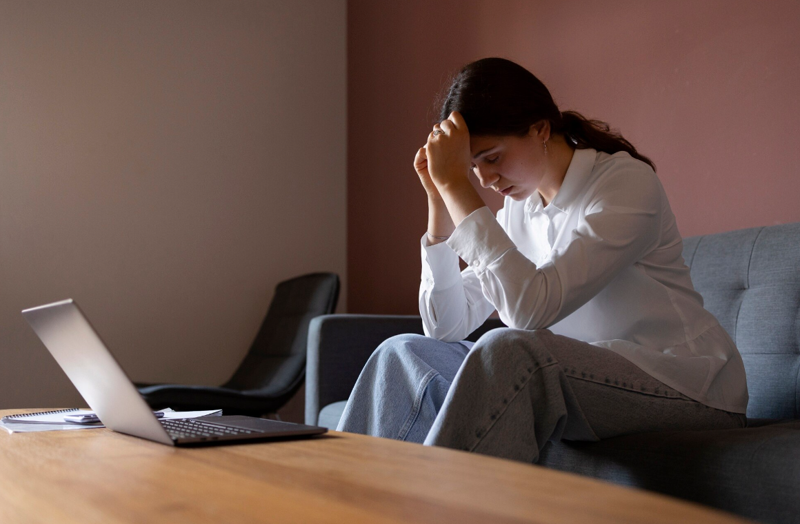 A woman sits on a couch leaning forward with her head in her hands, appearing stressed or overwhelmed while working on a laptop, representing hidden struggles of high-functioning addiction