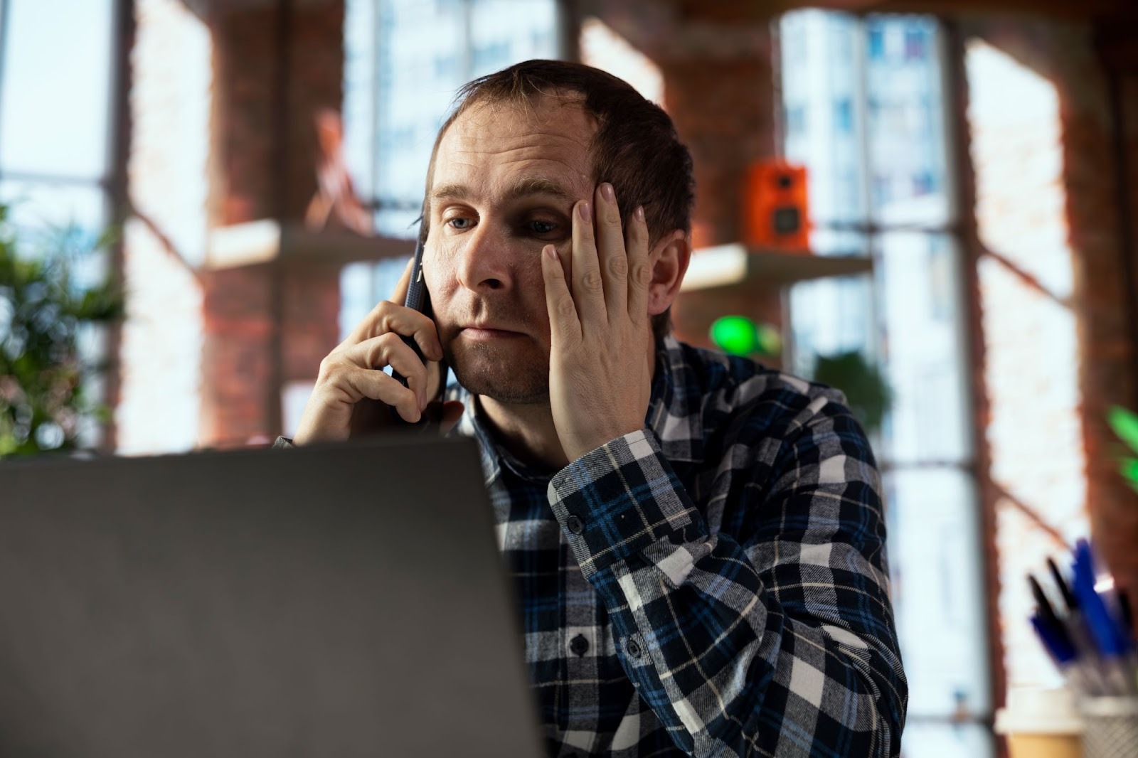 A man in a plaid top sitting in front of a laptop, making a phone call, and looking visibly stressed 