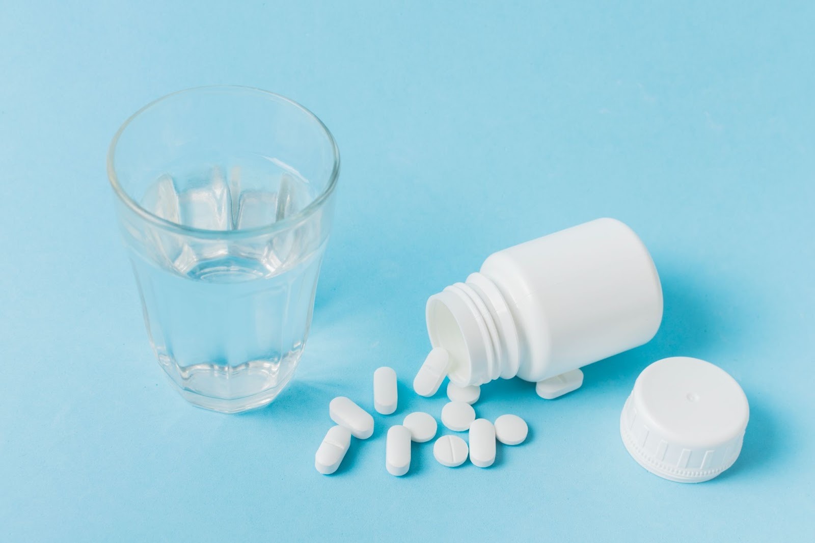 A glass of water sits next to an open white pill bottle on its side, spilling tablets onto a light blue surface, symbolizing Tylenol misuse