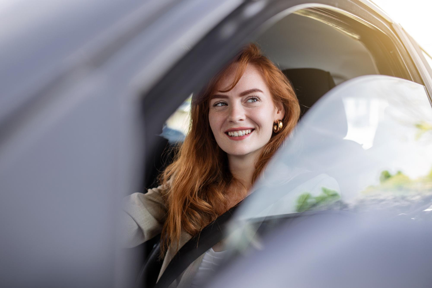 Smiling woman with red hair sitting in the driver’s seat of a car, looking out the window in natural daylight