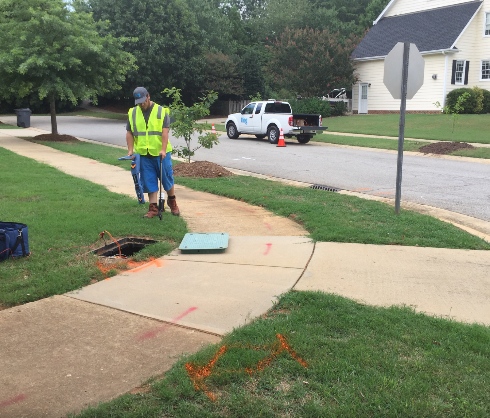 Ting fiber technician in front of a handhole where a fiber mainline is accessible. Public utilities have been marked with pain on the sidewalk and lawn.