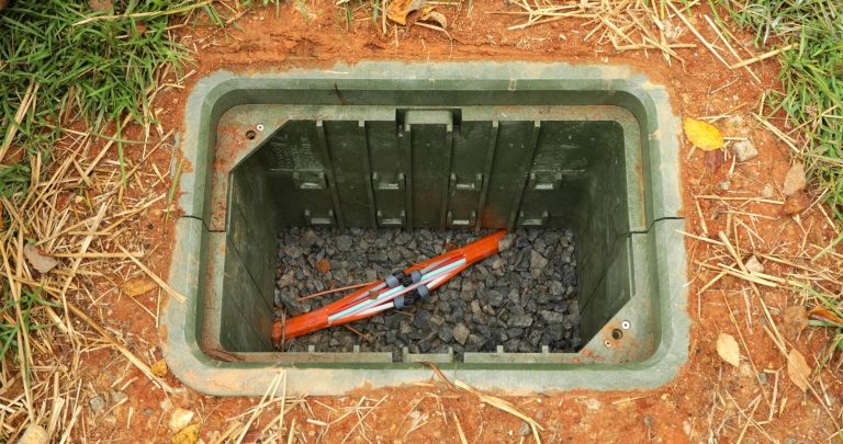 An example of an open green handhole flush with the grass and dirt ground. Inside are some red and white fiber internet cables, along with rocks for drainage.