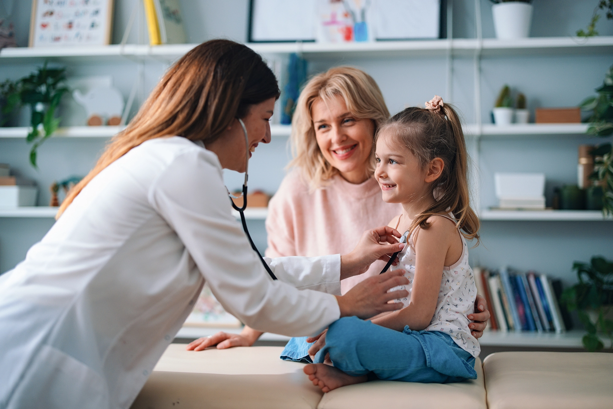 Female doctor uses stethoscope to check smiling young girl sitting on exam table with her mother.
