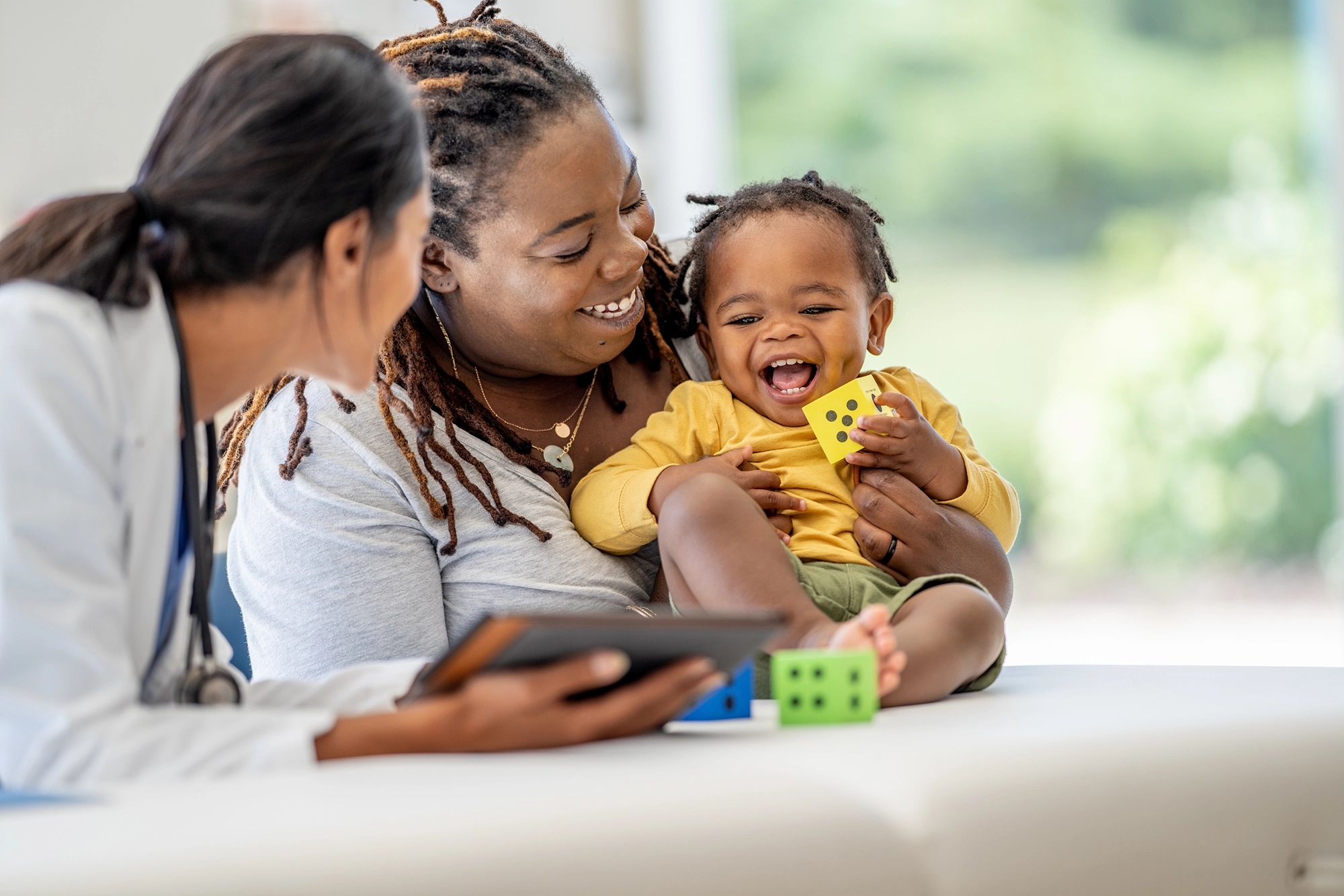 Smiling toddler held by their mother, interacting with doctor, playing with colorful blocks.