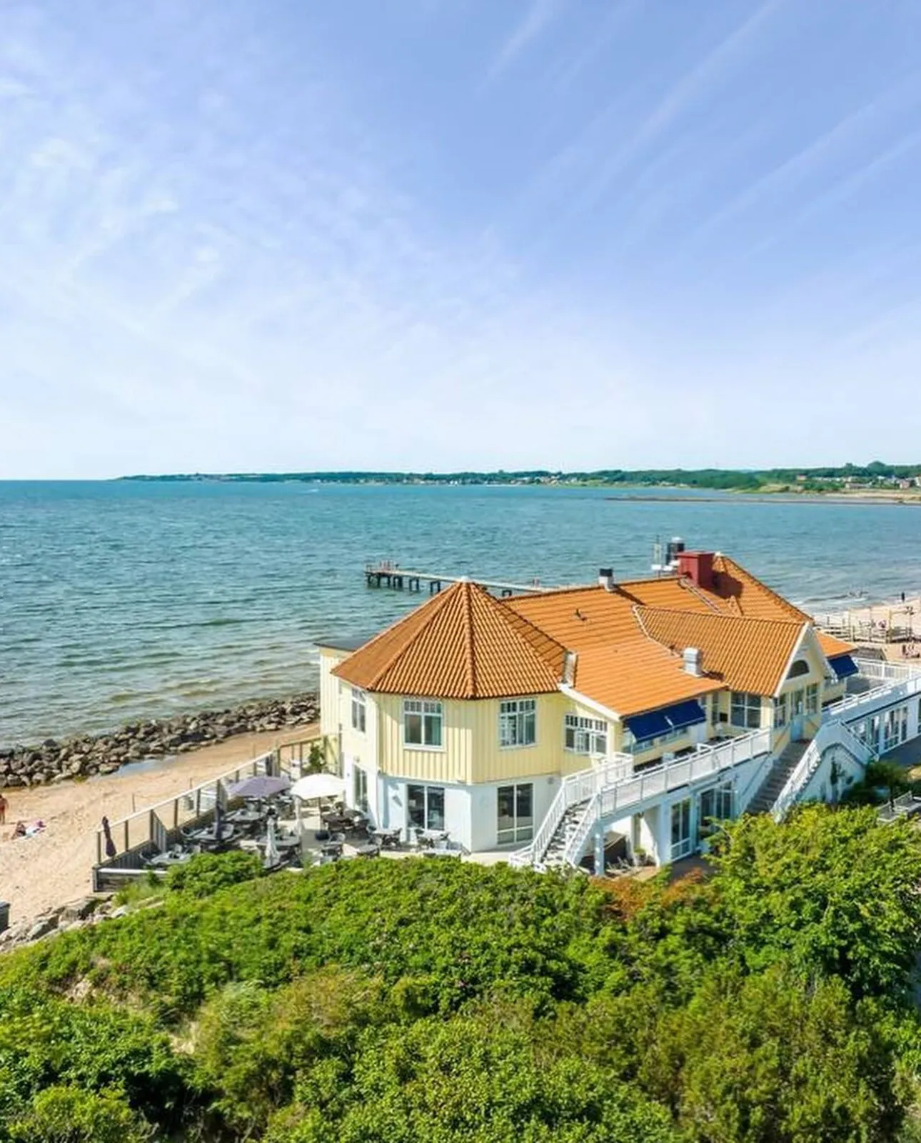 Gult hotell med oranget tak vid havet, omgivet av grönska och en strand med brygga i bakgrunden.