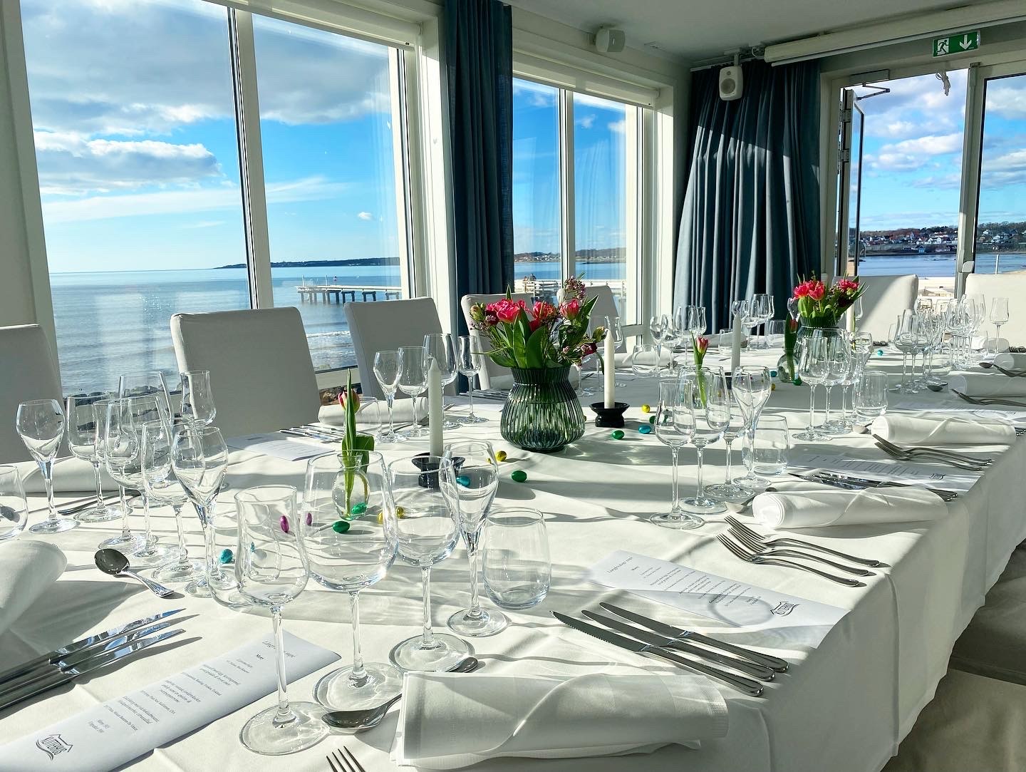 Påskdekorerat bord med glas, bestick och blommor vid sjöutsikt genom stora fönster.