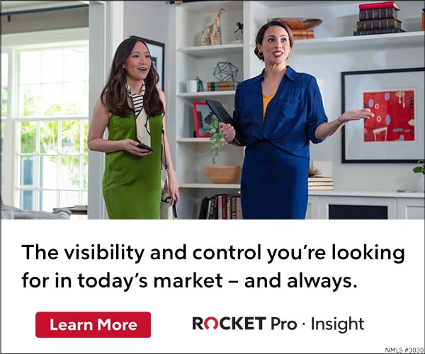 Real estate agent with female homebuyer standing indoors; one in a green dress and scarf, the other in a navy blue skirt and shirt holding a black folder.