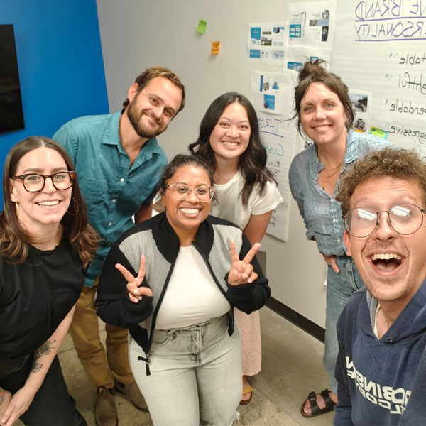 A "groupie" photo of five workshop participants and Rashmi Lopez in front of some workshop stickies on a wall