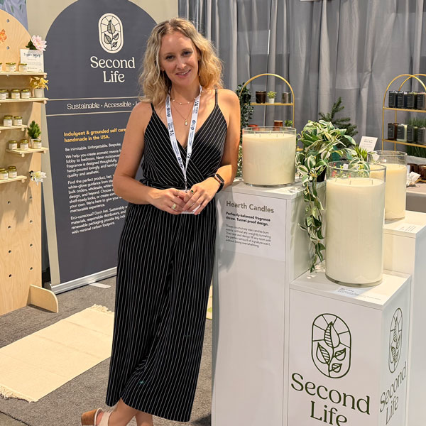 A woman stands in front of a tradeshow booth, leaning against three columns of different sizes holding large hearth candles and plants.