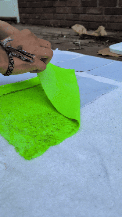 Hand peeling off a sheet of bright green paper from a white surface outdoors.