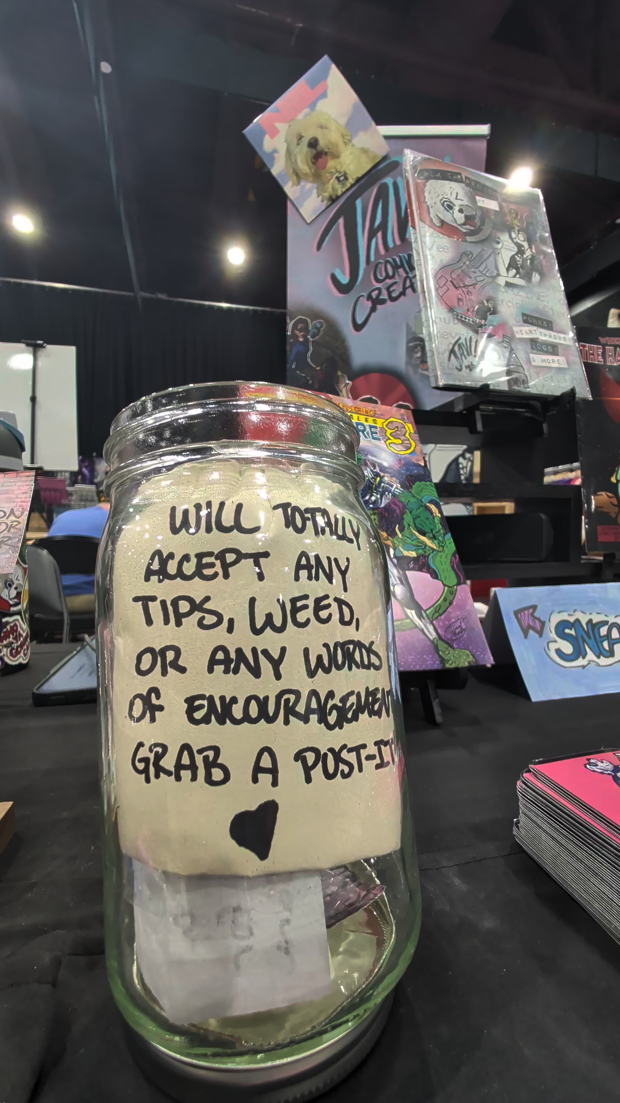 Glass jar with note saying tips, weed, or words of encouragement accepted, with booth art in background.