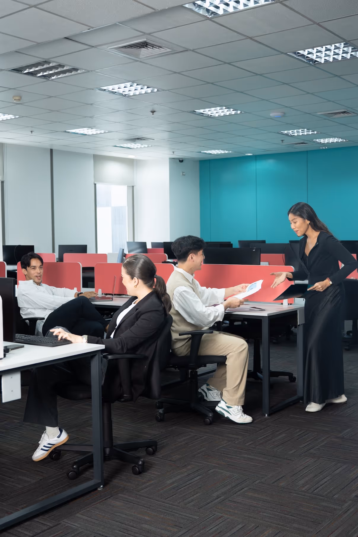 Four young professionals in an office, one woman standing handing papers to a seated man.