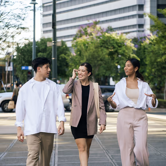Three young adults walking and talking on a city sidewalk with trees and buildings behind them.