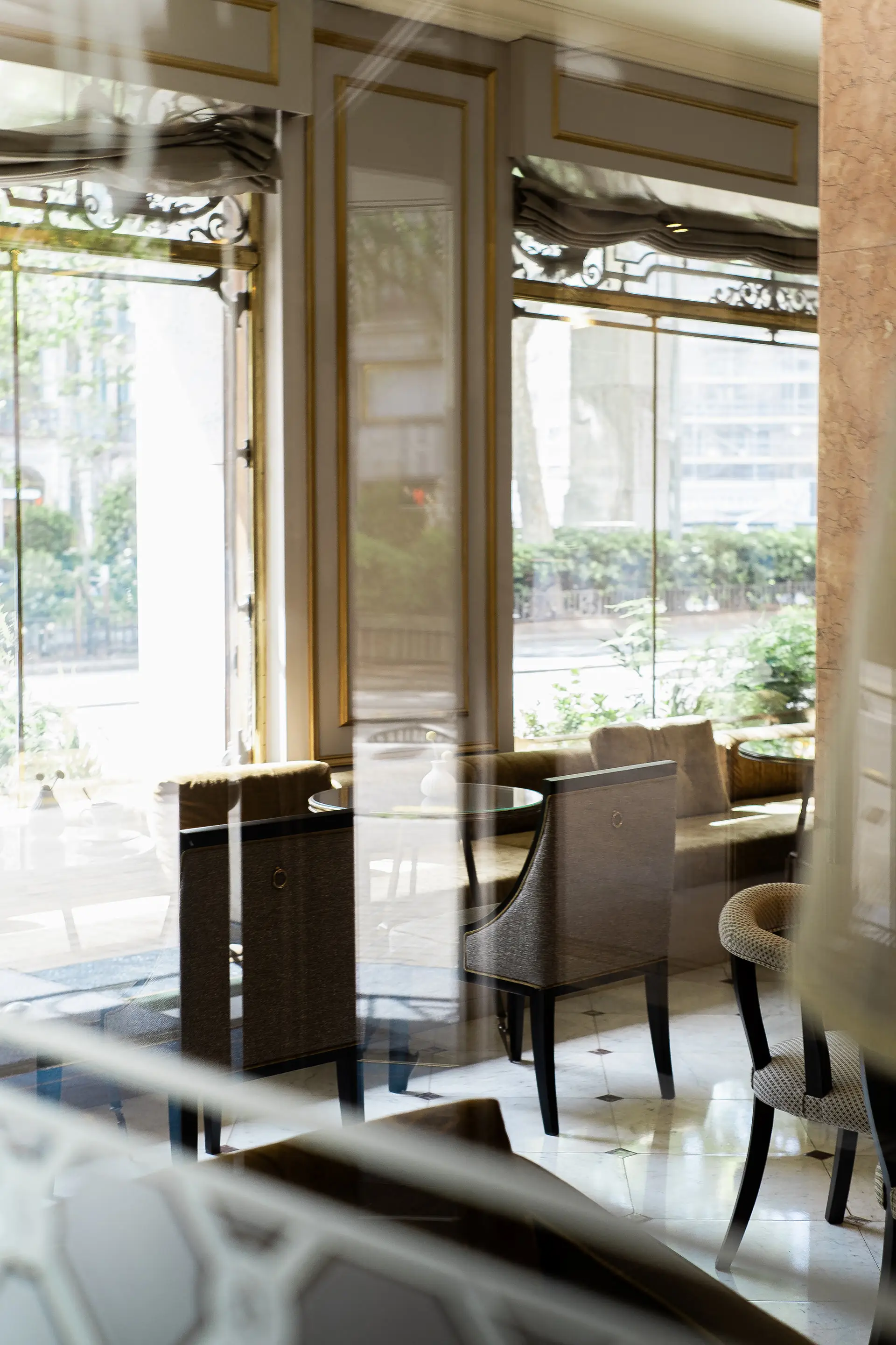 Una vista del interior al estilo café visto a través de grandes ventanales de vidrio, mostrando elegantes disposiciones de asientos con sillas acolchadas y mesas. El ambiente es luminoso, rodeado de vegetación y reflejos del paisaje urbano.