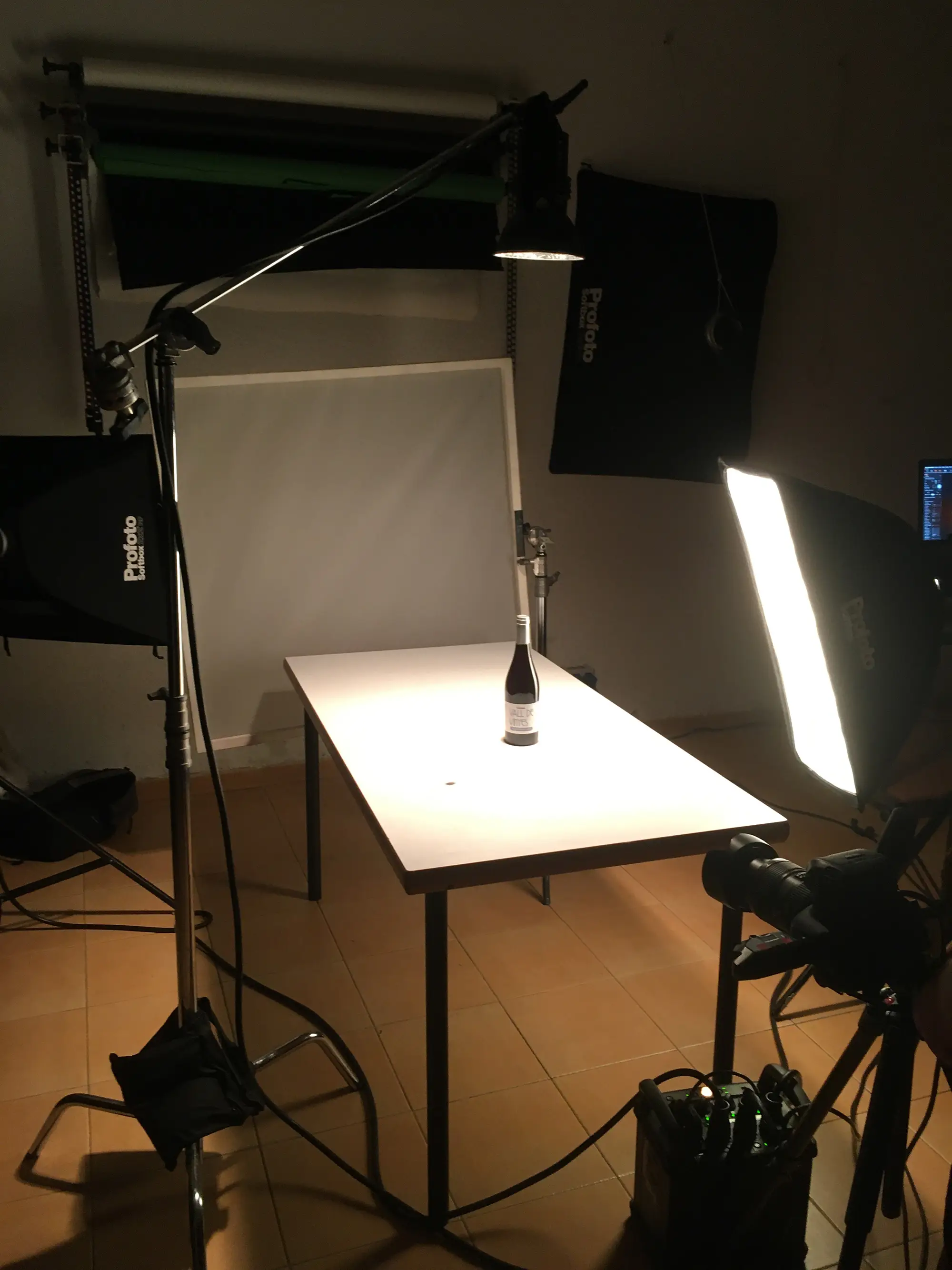 Un montaje de mesa en un estudio fotográfico con luces de softbox y una cámara posicionada para fotografiar una botella de vino colocada sobre una mesa blanca. La escena está iluminada para fotografía de producto, con un fondo minimalista e iluminación neutra.