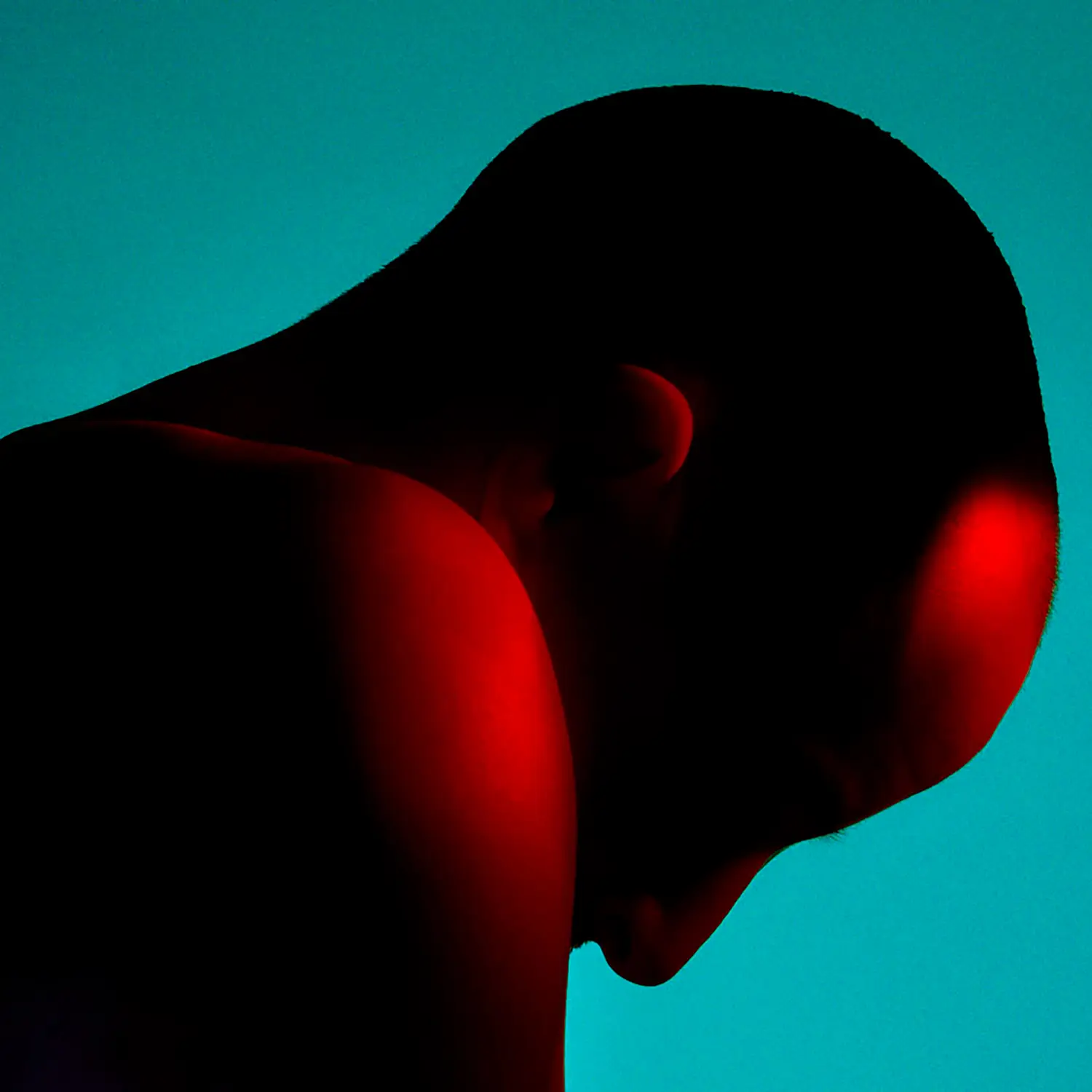 Un primer plano dramático del perfil de la cabeza y el hombro de una persona, iluminada con luz roja contra un fondo color teal. El contraste entre las sombras y los colores vibrantes crea un efecto visual impactante y atmosférico.