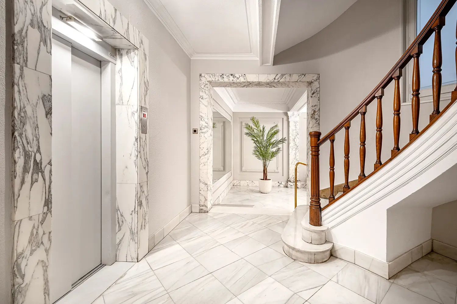 Entrada elegante con pisos y paredes de mármol, acompañada de una escalera clásica con barandillas de madera. Una palmera en maceta aporta un toque de verdor, mientras que las paredes espejadas amplifican el espacio, realzando su lujosa atmósfera.