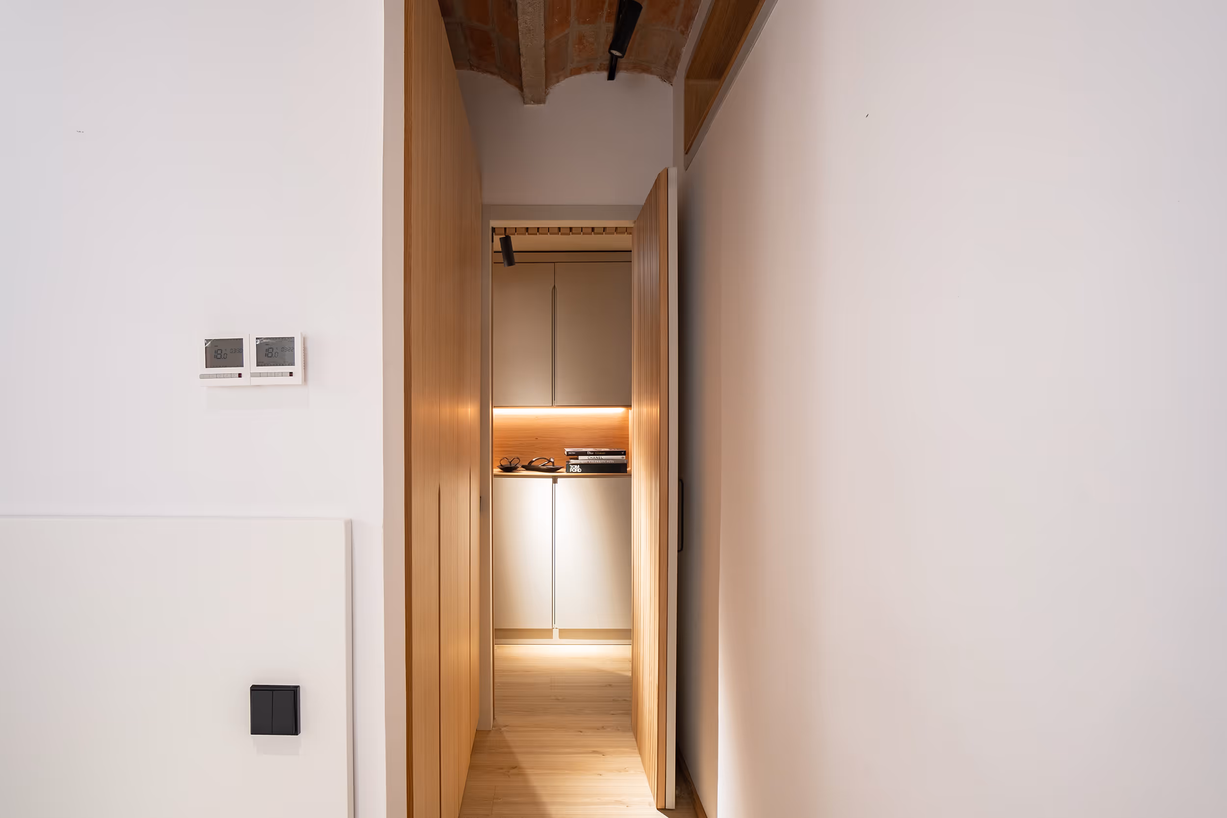 Un rincón acogedor con techo de madera y una luz suave que ilumina una zona de armarios con libros y vasos. El espacio compacto está enmarcado por puertas de madera