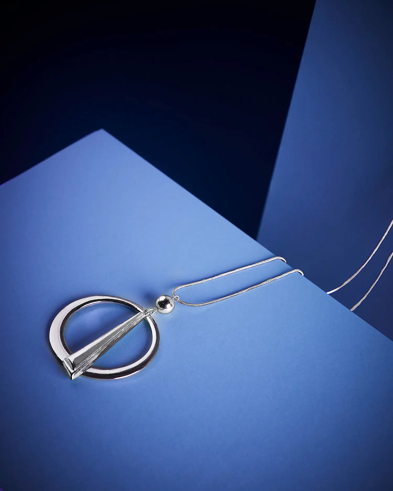 Un moderno collar plateado con un colgante geométrico compuesto por una barra triangular dentro de un marco circular. La joya está dispuesta sobre una superficie minimalista azul, con una iluminación suave que enfatiza su acabado pulido.