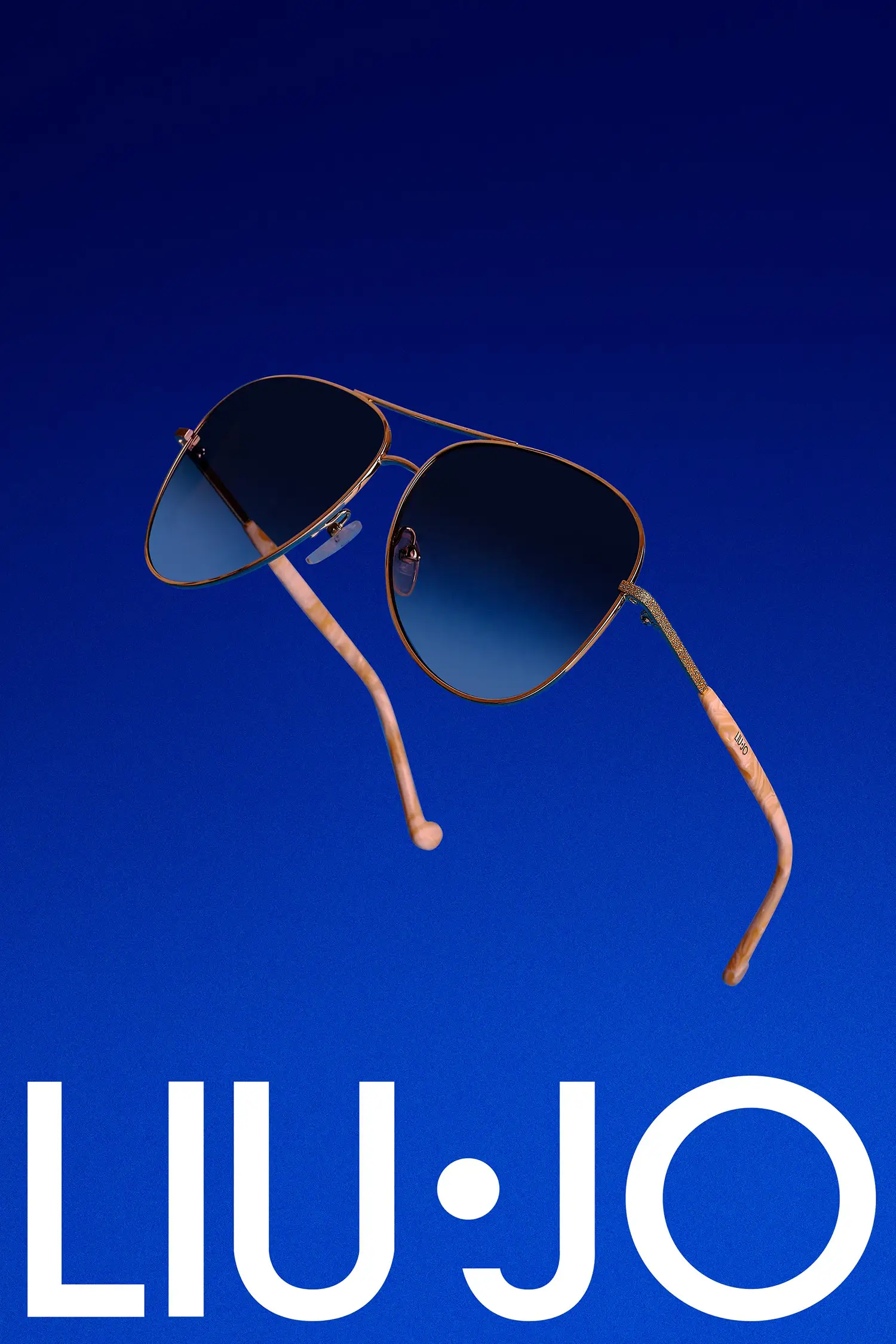 Still life Gafas de la marca Liu-jo con montura dorada y lentes en degradado azul suspendidas sobre un fondo azul sólido. Las patillas curvadas presentan una textura única marmoleada, destacando la elegante artesanía.tography of sunglasses in the air with blue background