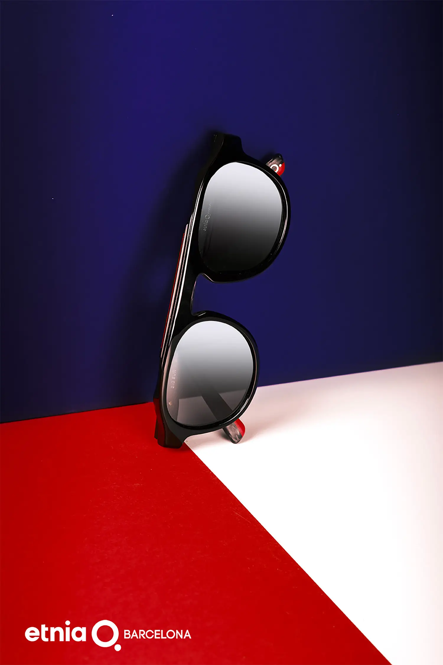 Unas gafas de sol de la marca Etnia redondas de montura negra con lentes espejados, apoyadas contra un audaz fondo de bloques de color en rojo, blanco y azul. Los reflejos en los lentes añaden profundidad a la composición estilizada.