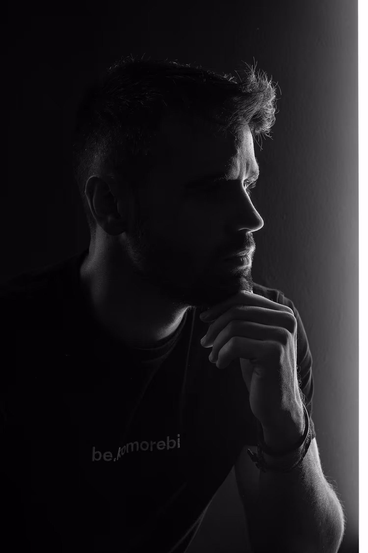 Retrato en blanco y negro de una persona de perfil, apoyando la barbilla en la mano en una pose reflexiva. Una iluminación sutil resalta los contornos de su rostro, mientras lleva una camisa con el logo "be.komorebi" parcialmente visible en el pecho.