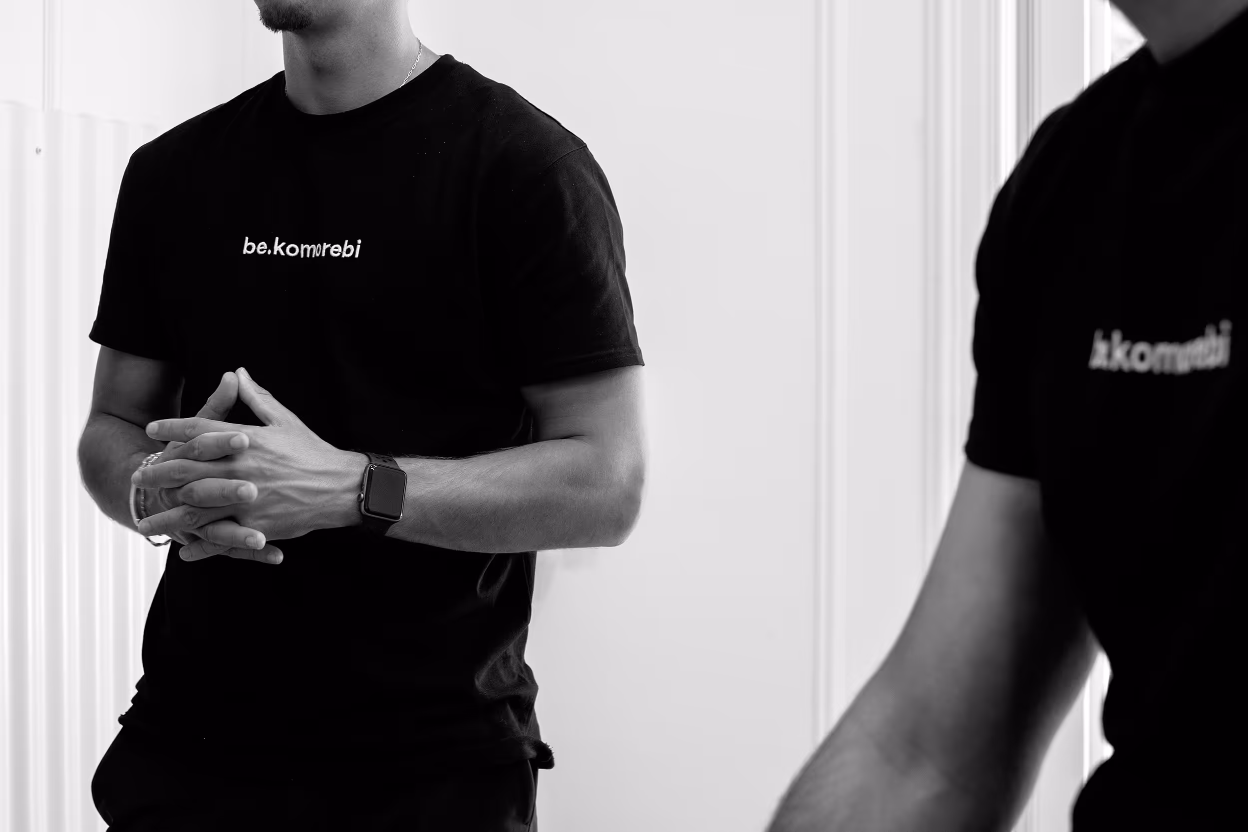 Primer plano en blanco y negro de dos personas vistiendo camisetas con el logo "be.komorebi" visible, con una de ellas juntando las manos frente al pecho. La palabra "Corporativo" en negrita y texto blanco se superpone a la imagen.