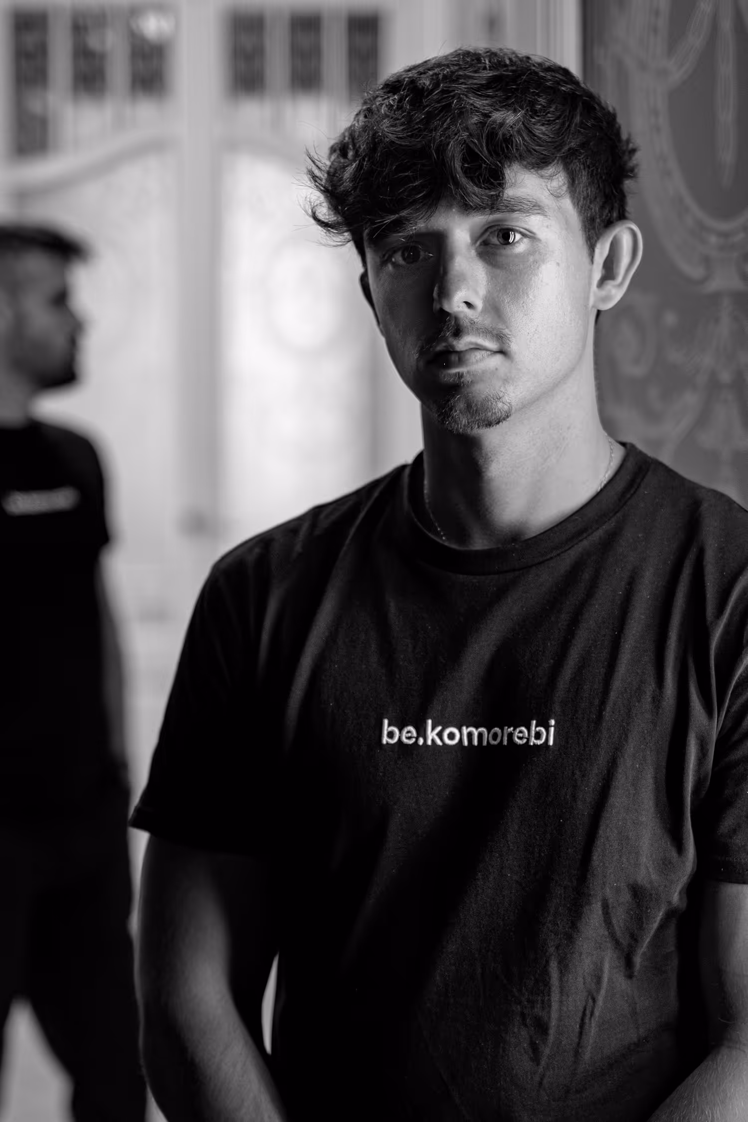 Retrato en blanco y negro de una persona vistiendo una camiseta "be.komorebi", posando enfocada con una expresión neutral. En el fondo, otra persona aparece ligeramente desenfocada, añadiendo profundidad a la composición contra un fondo suavemente texturizado.