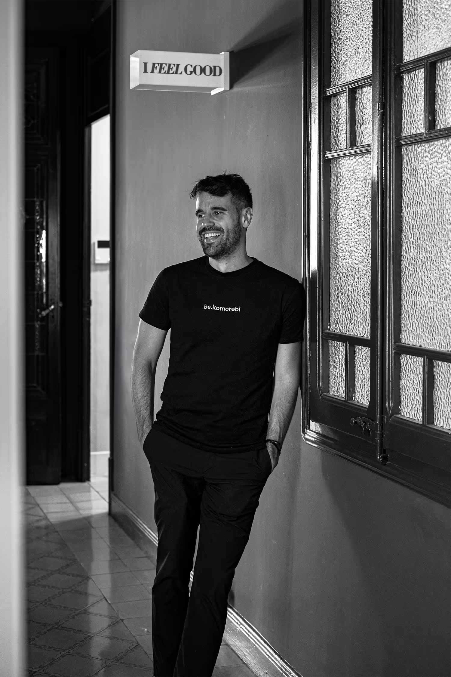Foto en blanco y negro de una persona sonriente apoyada contra una pared, vistiendo una camiseta "be.komorebi". Un letrero iluminado arriba dice "I FEEL GOOD," y ventanas de vidrio texturizado enmarcan parte del fondo, creando una atmósfera relajada y alegre.