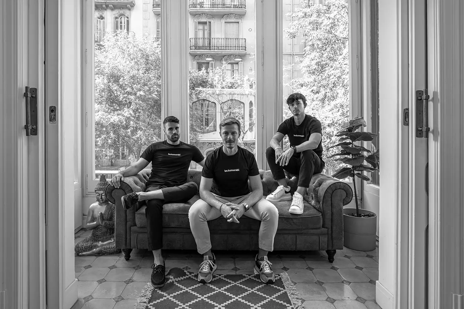 Tres personas sentadas de manera casual en un sofá en una habitación luminosa con grandes ventanas que dan a un exterior frondoso, cada una vistiendo camisetas "be.komorebi".
