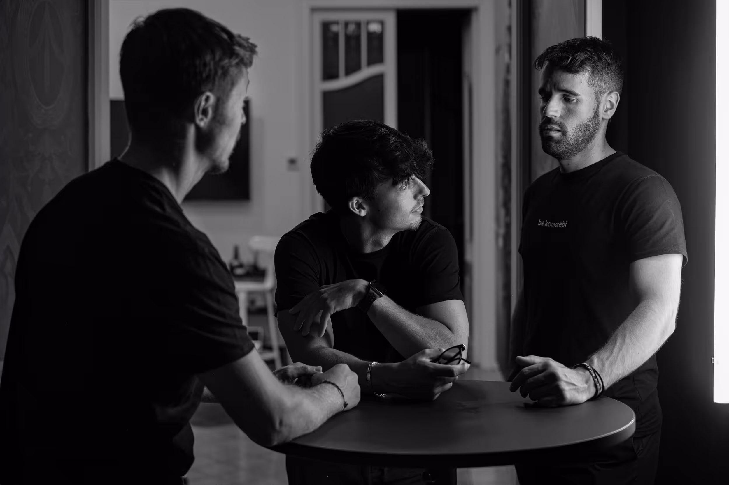 Un momento más íntimo mientras tres personas conversan alrededor de una mesa pequeña en un entorno interior tenuemente iluminado, con expresiones concentradas.