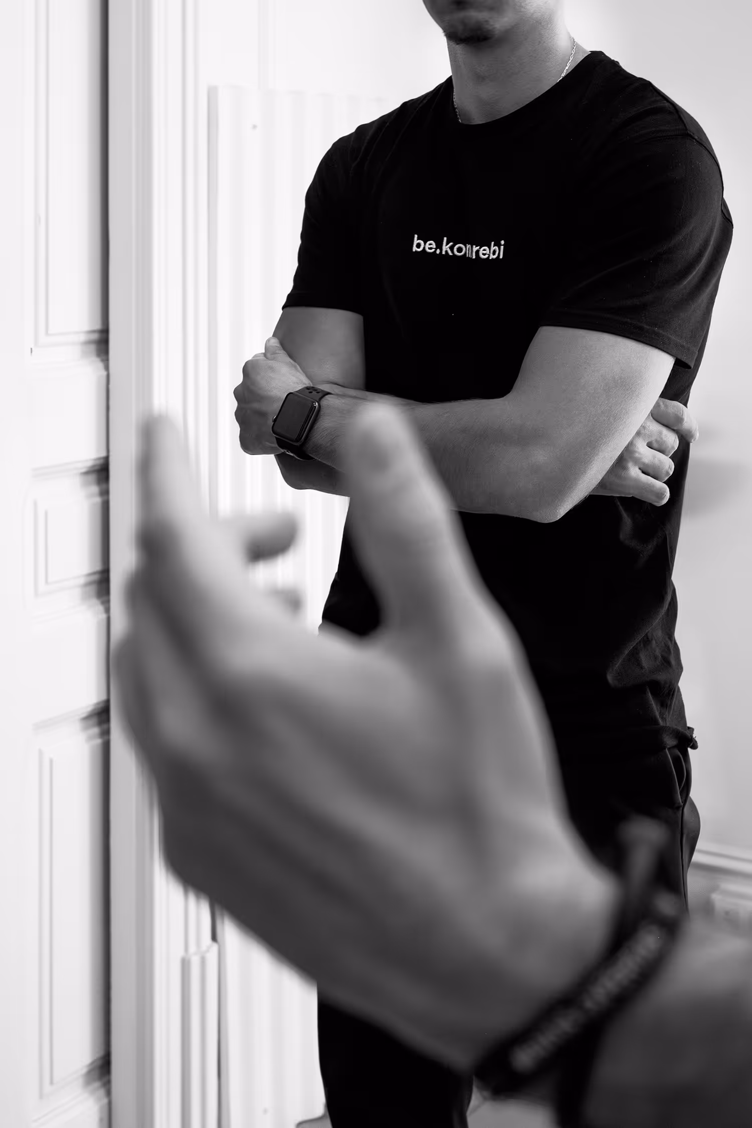 Fotografía en blanco y negro de dos personas conversando. En primer plano, una mano en gesto de diálogo desenfocada, mientras al fondo se observa a una persona con los brazos cruzados, vistiendo una camiseta negra con un logo blanco y un reloj digital en la muñeca.