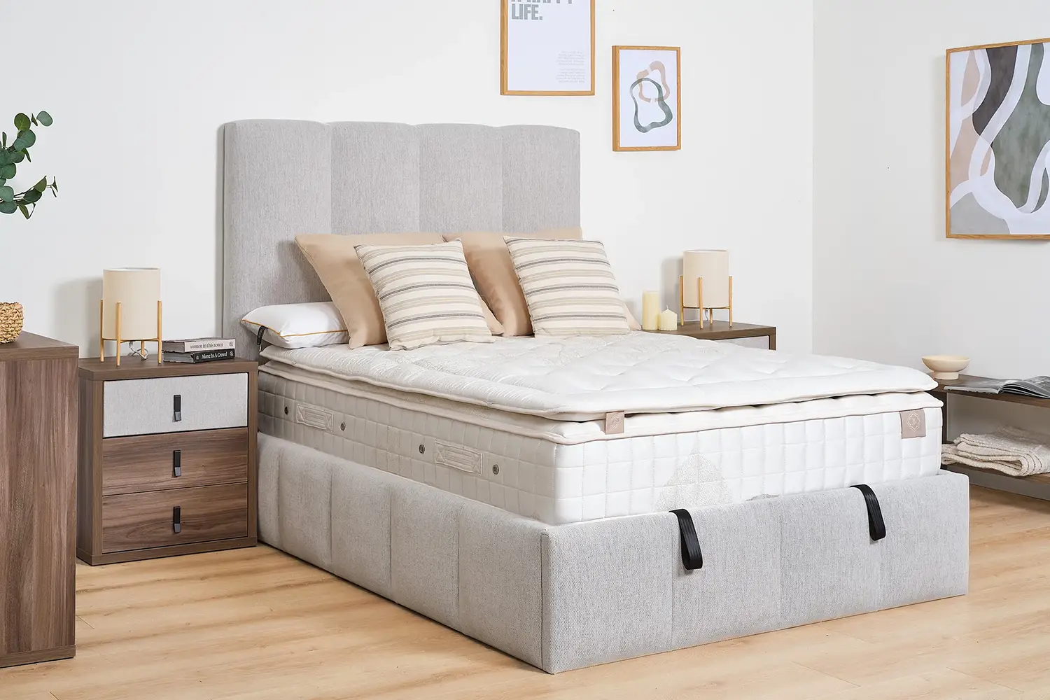 Cama doble con cabecero tapizado en gris claro y colchón de muelles ensacado, decorada con cojines en tonos neutros. Dormitorio moderno con muebles de madera, lámparas de mesa y cuadros minimalistas en la pared.