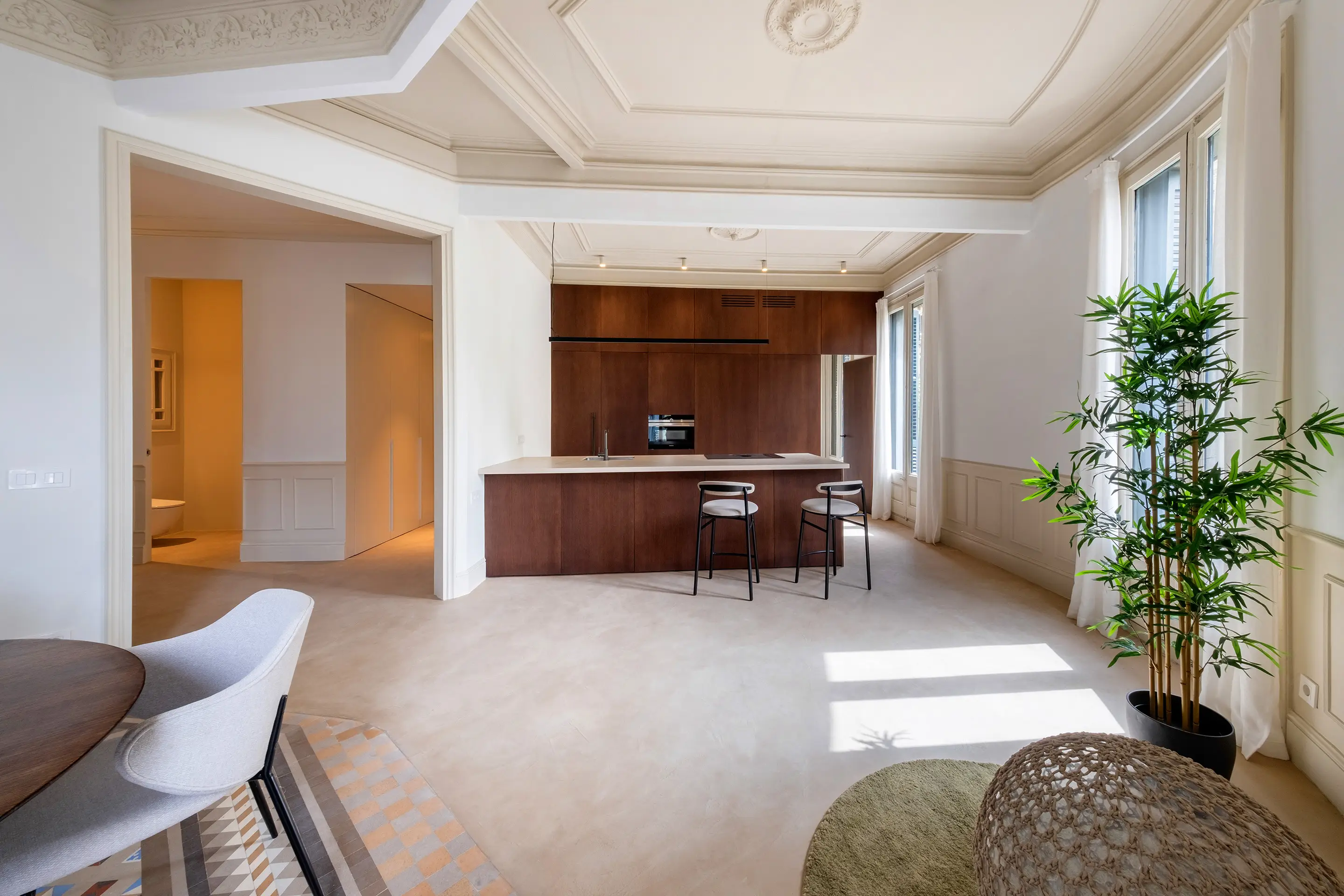 Una amplia cocina con una isla de madera oscura y dos taburetes, situada frente a un fondo de grandes ventanas que permiten la entrada de luz natural. La habitación cuenta con detalles ornamentados en el techo, paredes de colores claros y un diseño minimalista con acentos modernos.