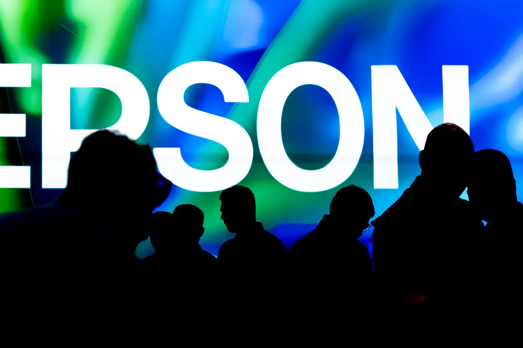 Asistentes en silueta frente a una pantalla de Epson durante una feria tecnológica internacional, fotografía profesional de evento corporativo.