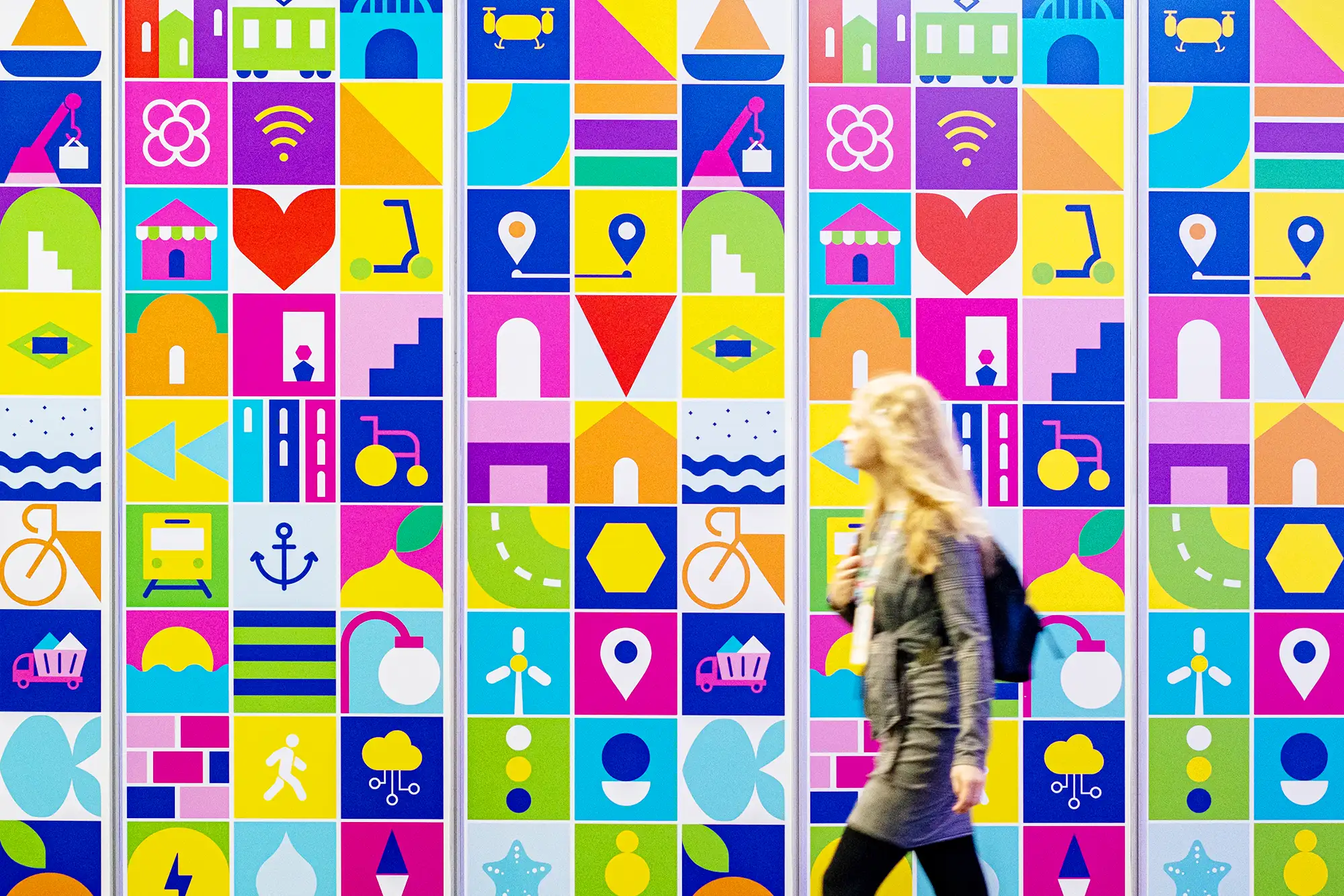 Asistente caminando frente a un mural gráfico colorido durante una feria internacional, fotografía creativa de evento corporativo.