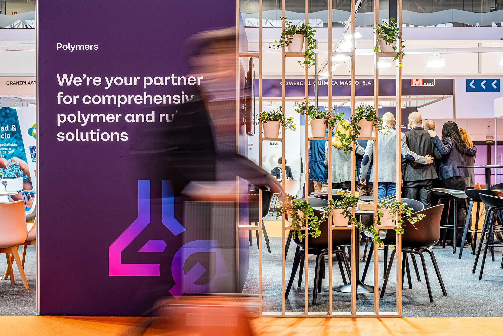Stand de feria con panel morado de empresa de polímeros, persona en movimiento desenfocada en primer plano y zona de reunión con mesas, sillas y plantas decorativas al fondo.