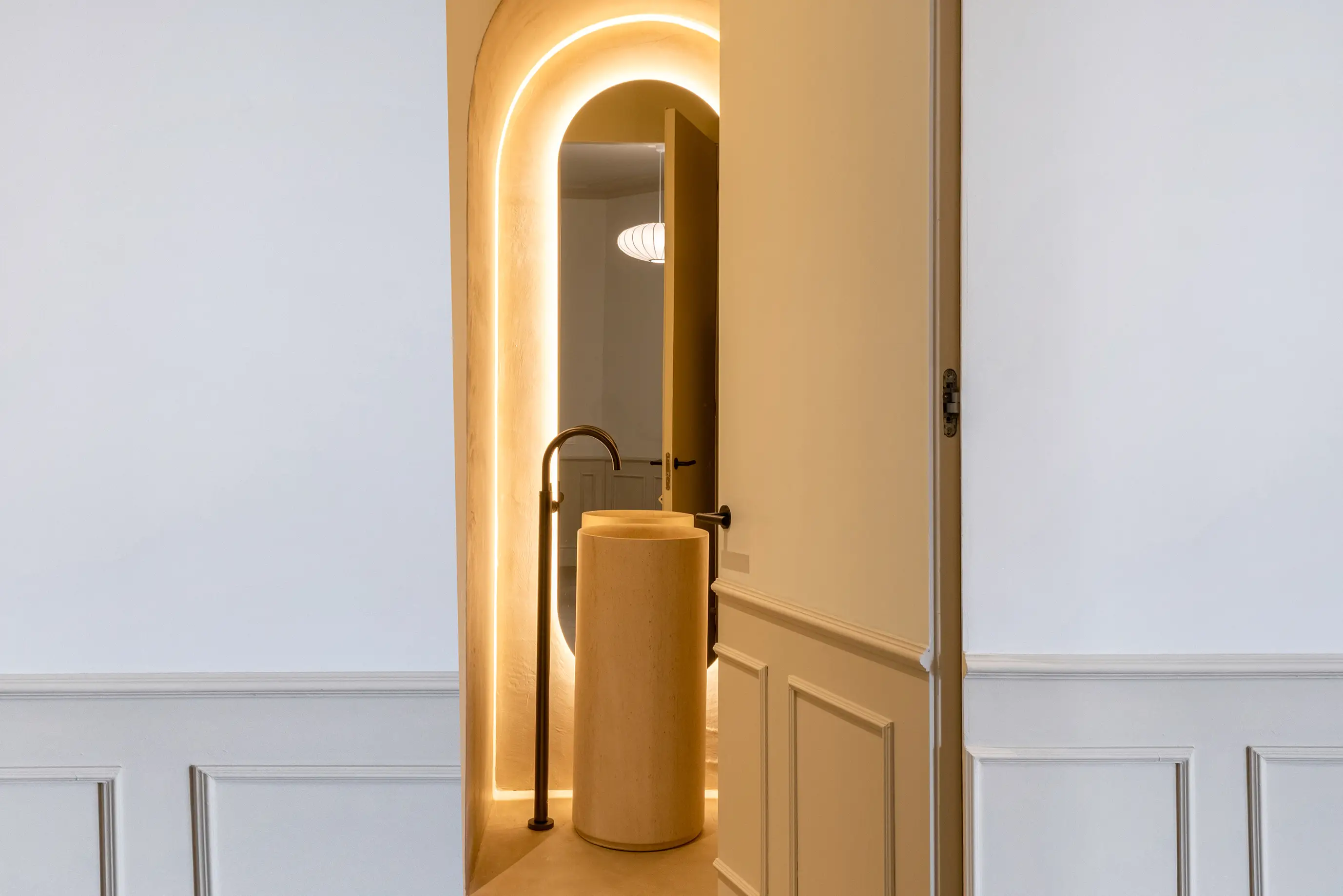 Interior contemporáneo con lavabo de piedra cilíndrico y grifo negro mate, enmarcado por un arco iluminado con luz cálida indirecta. Fotografía de interiorismo con tonos neutros y ambiente elegante.