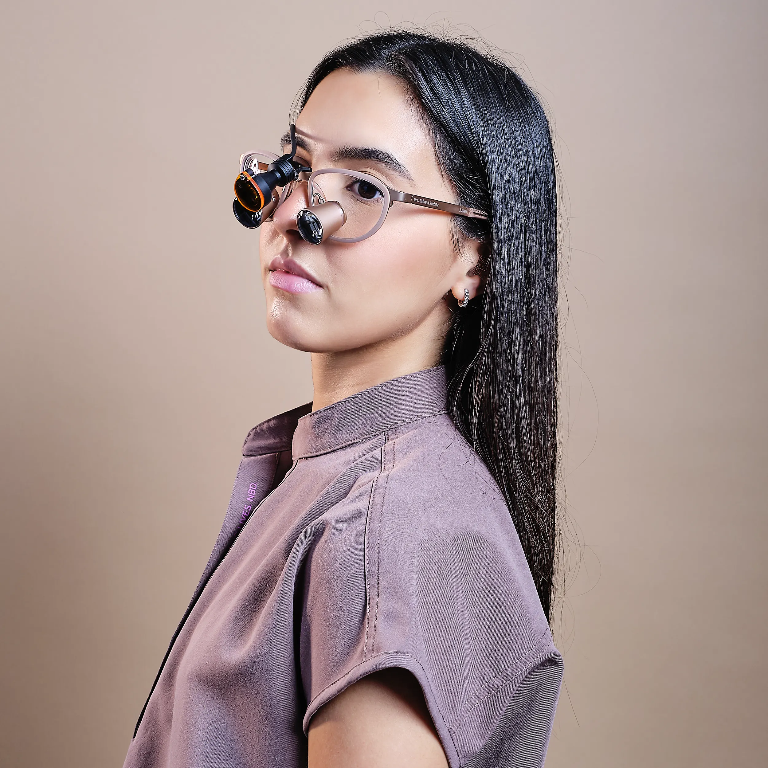 Side profile studio portrait of a woman with long dark hair wearing translucent glasses, photographed against a soft beige background.