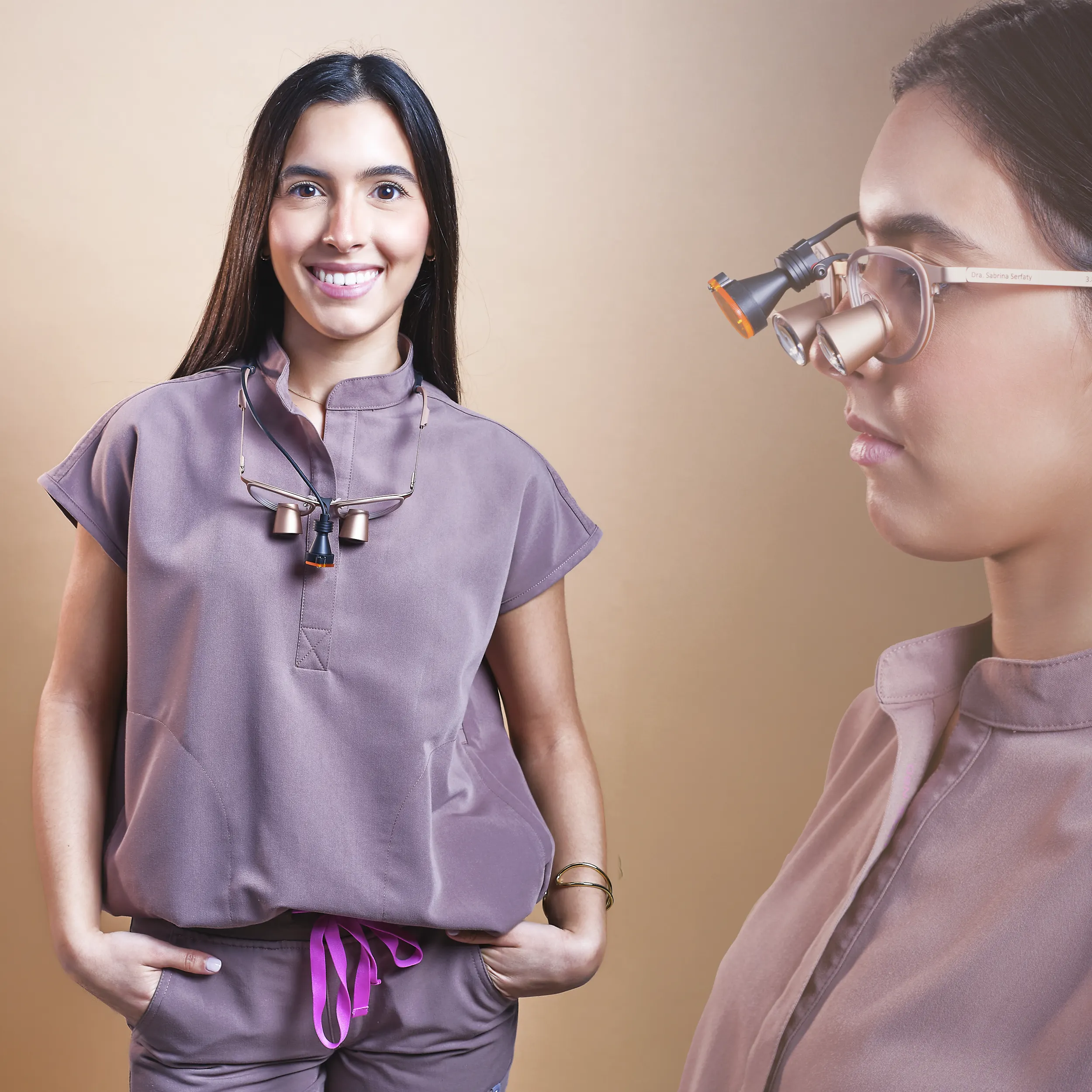 Studio portrait of a smiling woman wearing a casual short-sleeve top and glasses, standing relaxed against a warm neutral background.