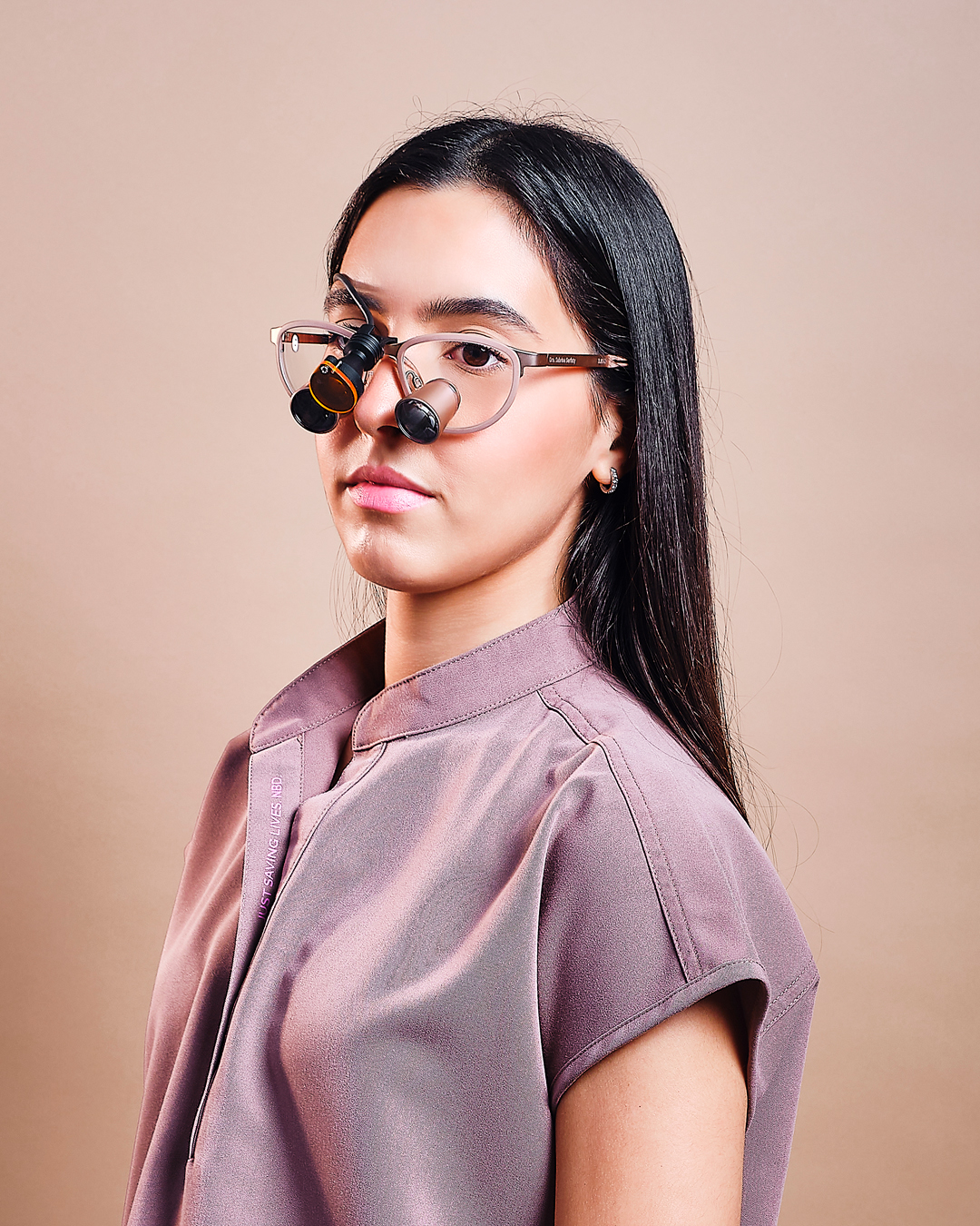Studio portrait of a woman with long black hair wearing translucent glasses, photographed against a soft beige background.